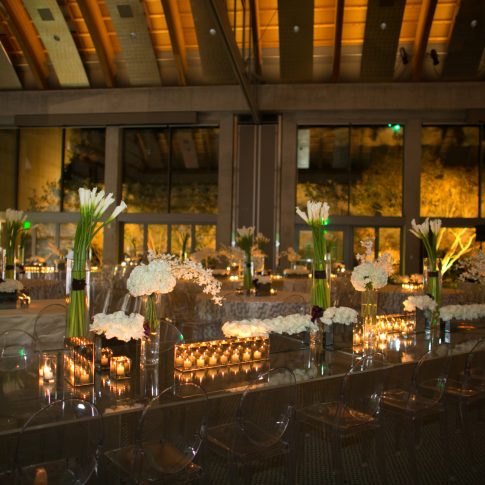 Elegant private event venue with mirrored dinner tables, clear ghost chairs, white floral arrangements, tall calla lilies, and abundant candlelight.