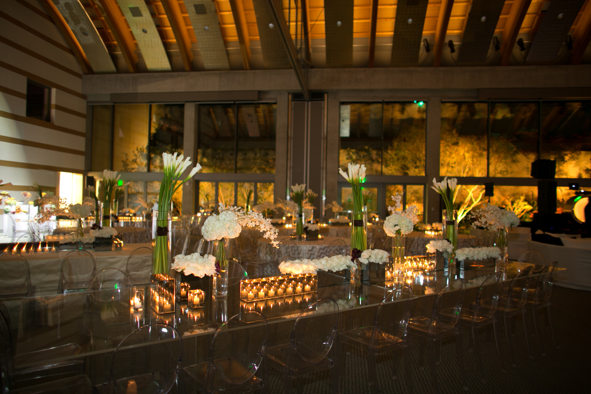 Elegant private event venue with mirrored dinner tables, clear ghost chairs, white floral arrangements, tall calla lilies, and abundant candlelight.