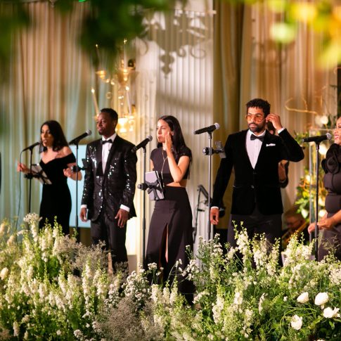 Live band performing at an elegant private hotel event with formal black attire, lush white and green florals, and warm ambient lighting.