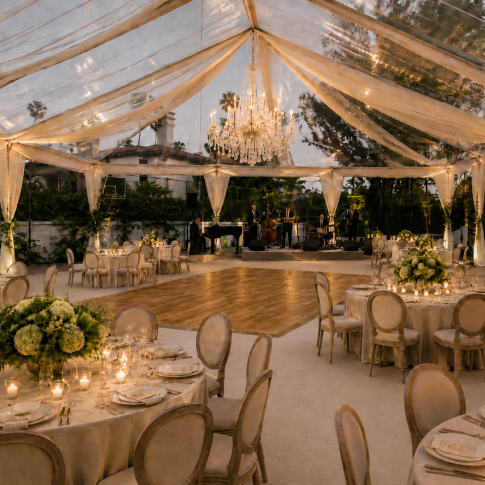 Elegant private estate wedding reception under a clear tent with ivory draping, crystal chandelier, warm wood dance floor, cream linens, candlelight, green and white florals, and a live band.