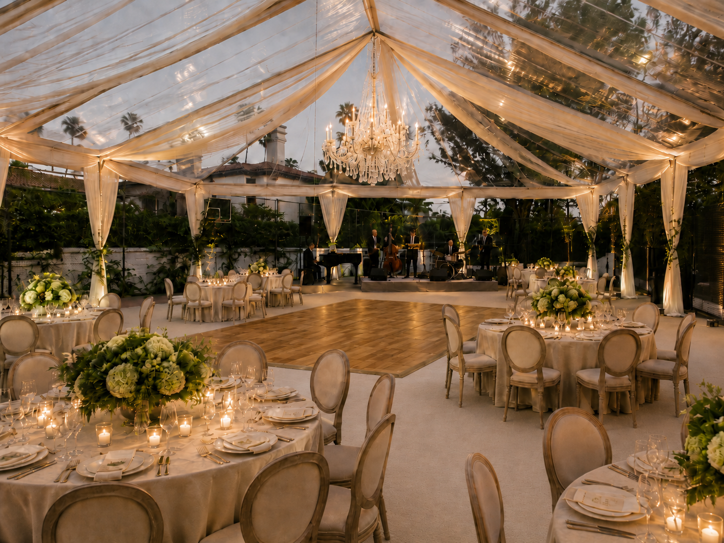 Elegant private estate wedding reception under a clear tent with ivory draping, crystal chandelier, warm wood dance floor, cream linens, candlelight, green and white florals, and a live band.