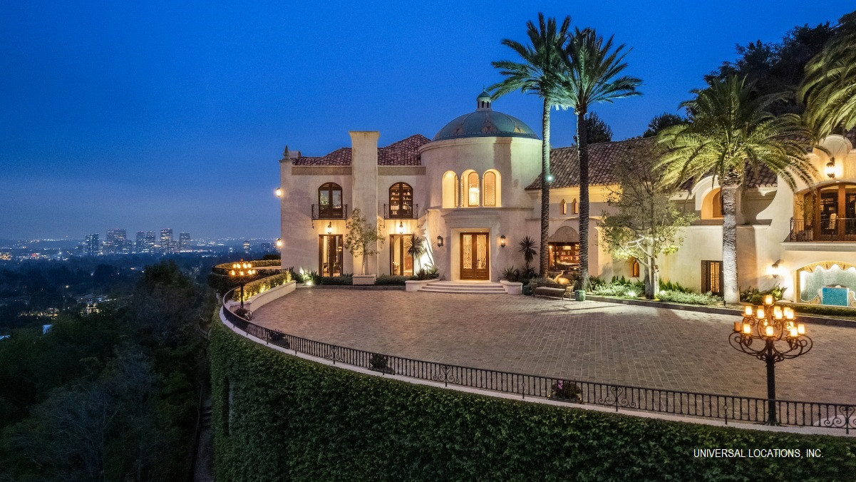 Grand illuminated motor court with Mediterranean architecture and sweeping Los Angeles skyline views at twilight.