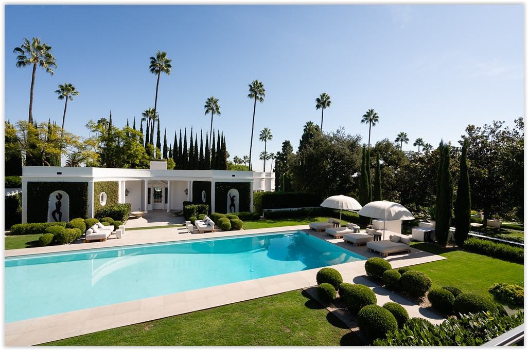 Resort-style pool terrace with luxury lounge seating, manicured hedges, and towering palm trees at a Beverly Hills estate.