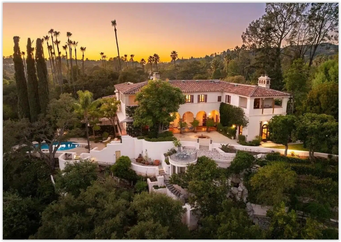 Sunset aerial view of a luxury Spanish Colonial estate surrounded by mature trees and hillside views.