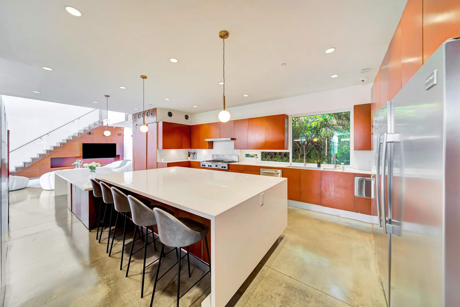 Sleek modern kitchen with large island, bar seating, stainless appliances, and abundant natural light from surrounding windows.