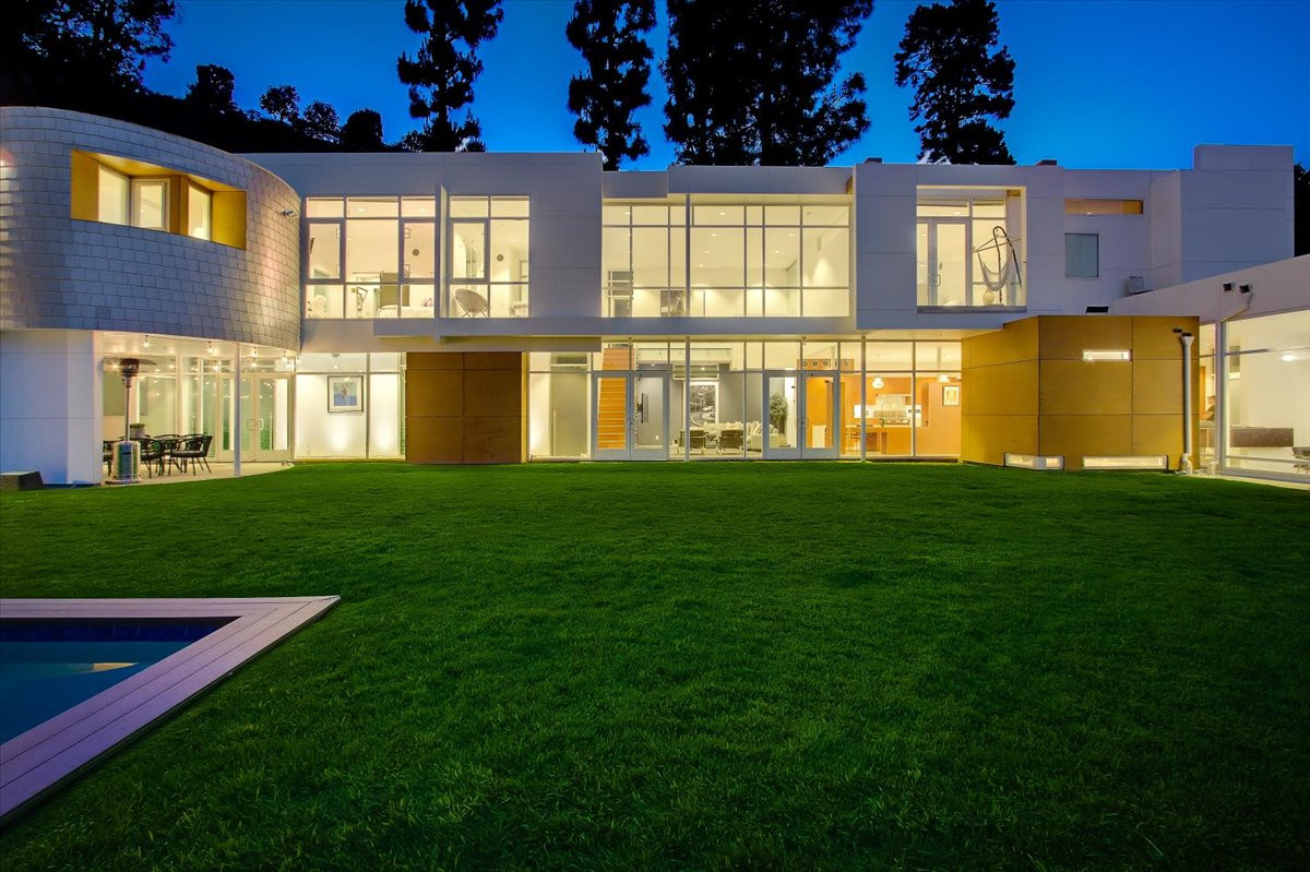 Modern multi-level estate exterior illuminated at night with expansive glass walls and green lawn overlooking canyon landscape.