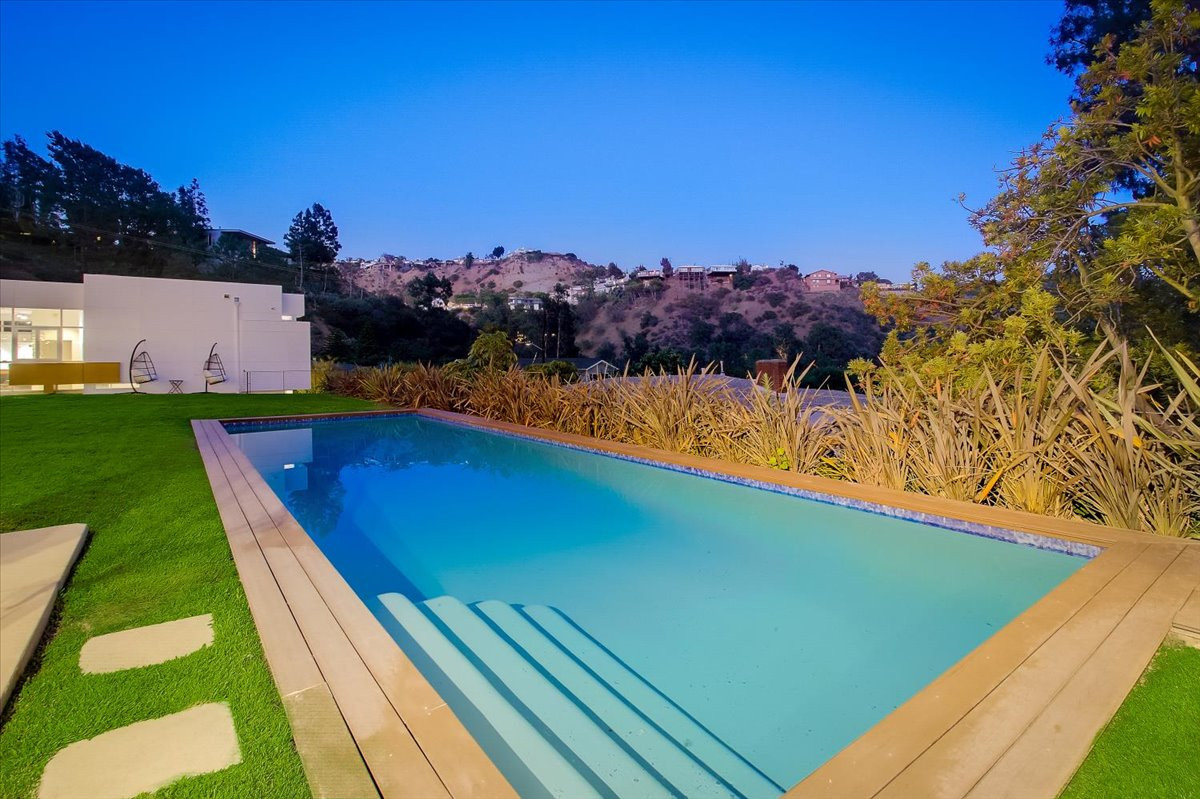 Rectangular swimming pool beside manicured lawn with lounge seating and hillside views at dusk.